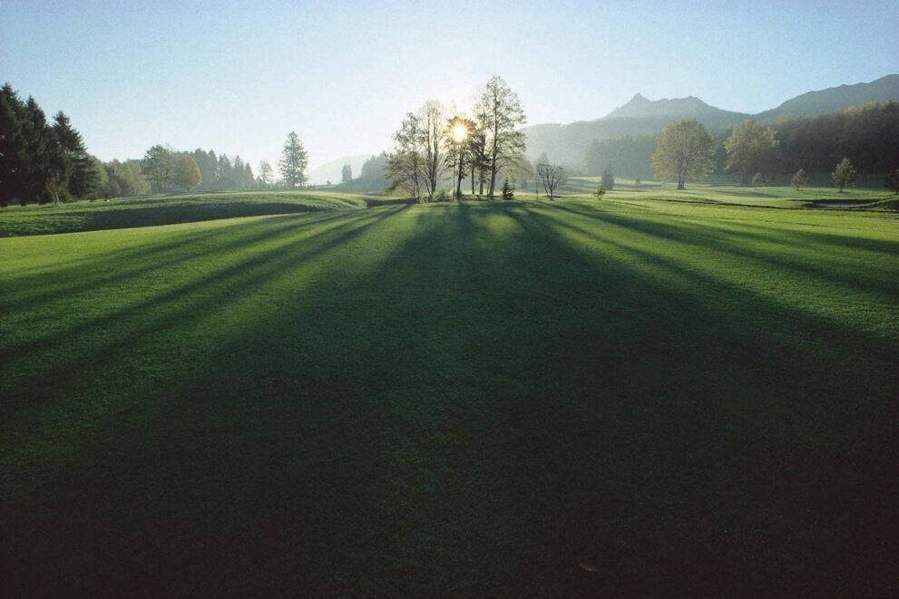 Ein grüner Golfplatz im Salzkammergut und hinter ein paar Bäumen geht die Sonne unter