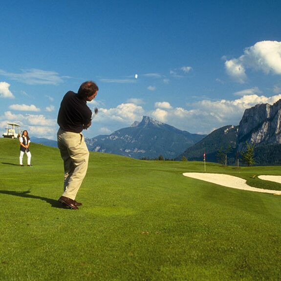 Ein Man schlägt den Golfball ab und zwei Frauen sehen ihn zu